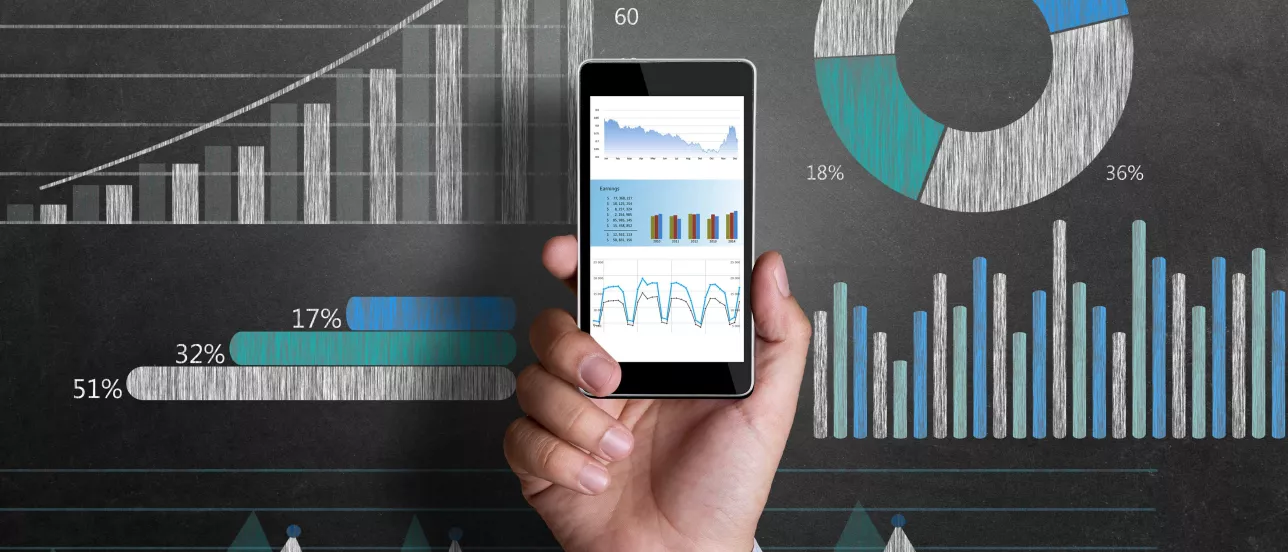 A hand holding a phone with graphs on the screen and in the background