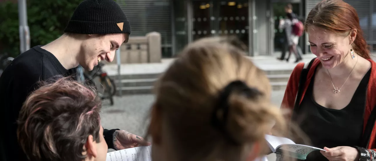 Four students laughing while holding and looking at books together in front of the LUX building