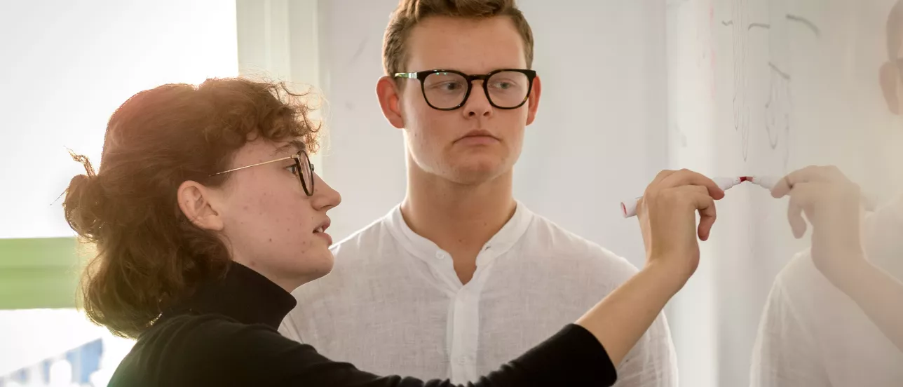 A student writing something on a whiteboard while another student is watching