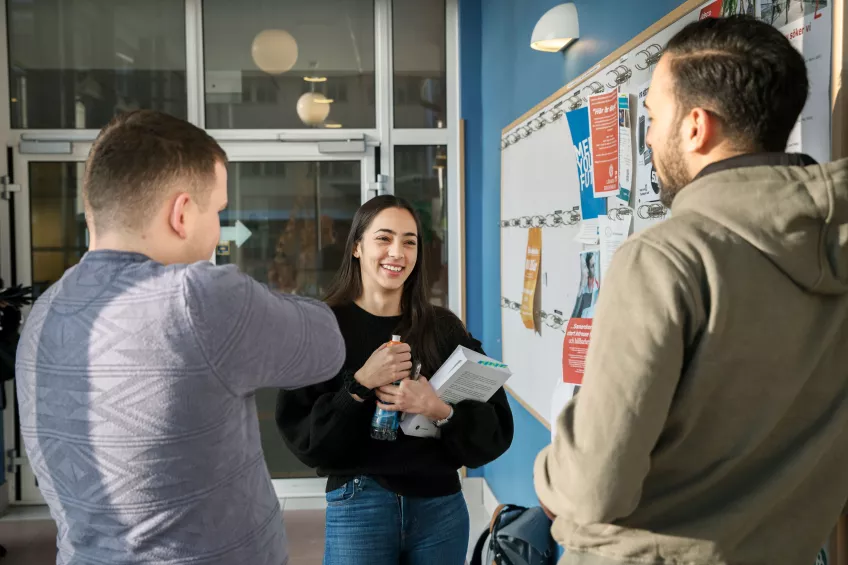 Three students talking to each other in front of a message board with flyers on it at Campus Helsingborg