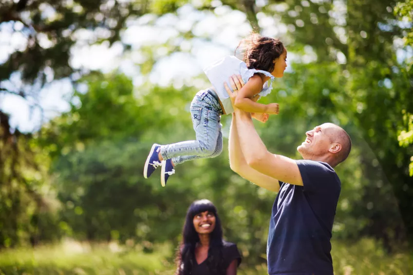 A father lifting up his child while the mum is watching them smiling