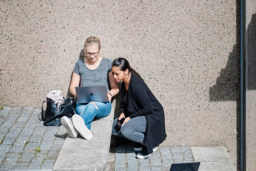 Two students seated against a wall and looking at a laptop outside